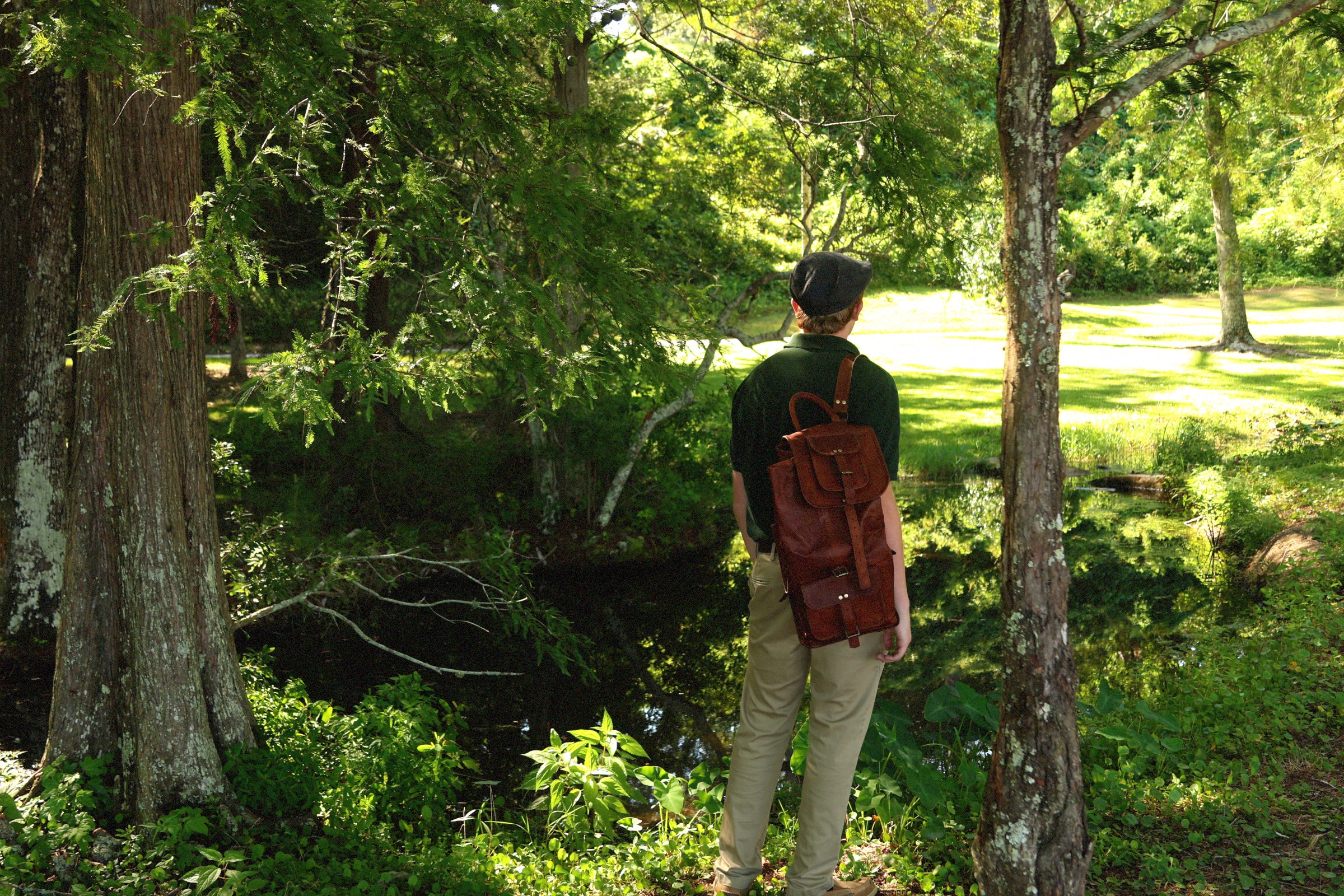 Model with brown leather rucksack facing a pond in a wooded area
