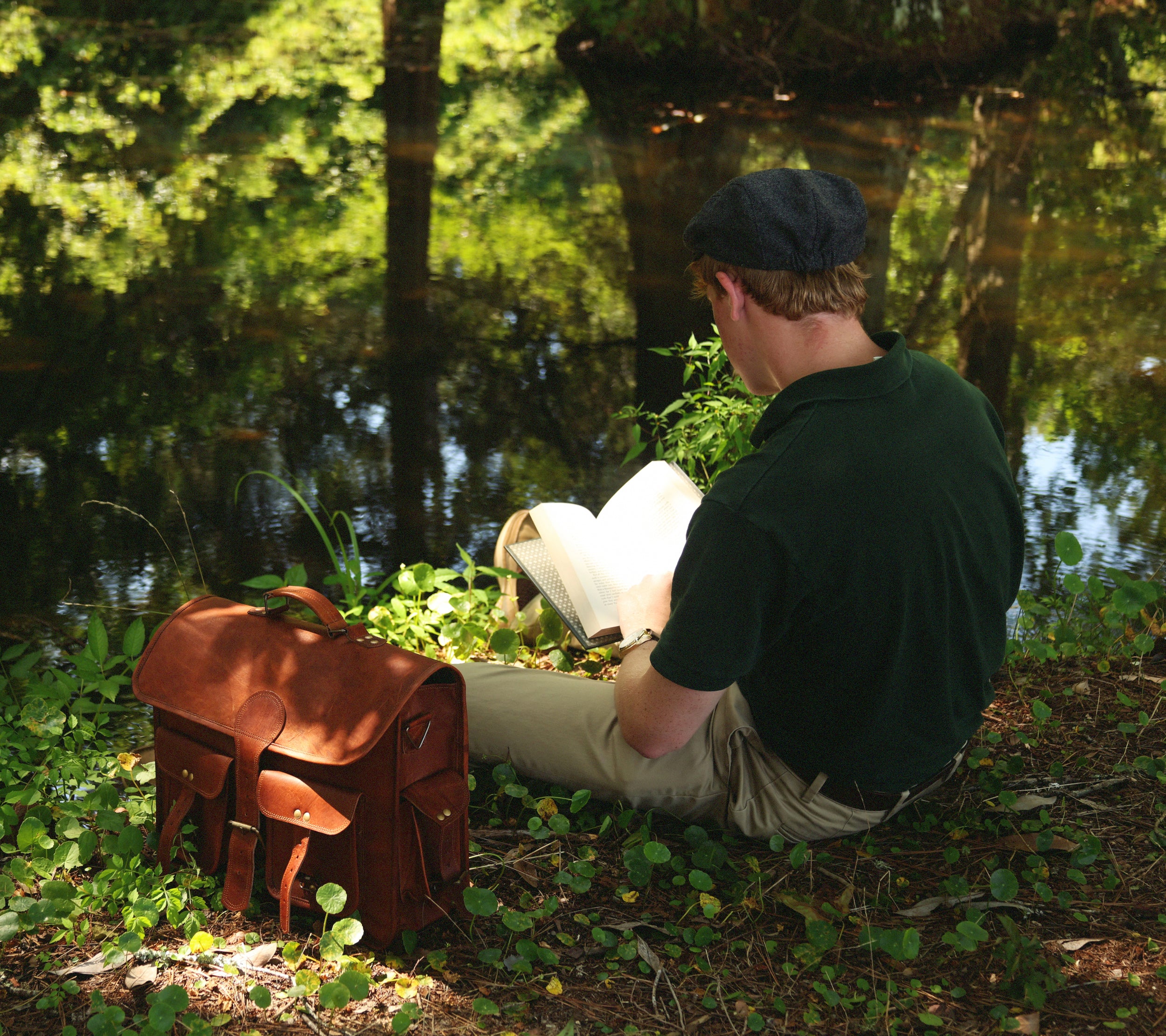 Model sitting next to a pond reading Robinson Crusoe with a satchel 